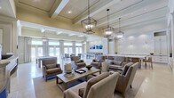 This refined single-floor amenity space is designed with a cohesive, upscale aesthetic that blends transitional architecture with comfortable, hospitality-inspired interiors. The open layout is anchored by soaring coffered ceilings adorned with striking geometric lantern-style pendant fixtures that cast warm, inviting light throughout. Polished travertine flooring runs continuously across the space, enhancing its sense of openness and grandeur.

The main lounge area features multiple groupings of tailored upholstered armchairs and sofas in neutral tones, centered around a wood coffee table, creating an ideal setting for relaxed gatherings. A classic fireplace with a refined mantel serves as a focal point in an adjacent seating area, flanked by French doors that invite abundant natural light. Framed artwork and built-in wall sconces add curated detail to the walls.

The game room accommodates a full-sized billiard table alongside additional gaming surfaces, all bathed in natural light from a wall of French doors and floor-to-ceiling drapery panels. A dedicated catering kitchen showcases floor-to-ceiling white cabinetry, marble countertops, a decorative patterned tile backsplash, a large central island with barstool seating, and a coffee station, making it highly functional for events and social gatherings.