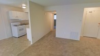 This single-floor residential space offers a practical and functional layout designed for comfortable everyday living. The main living area is open and spacious, featuring neutral-toned wall-to-wall carpeting that adds warmth underfoot. Soft, even lighting from a ceiling-mounted fixture illuminates the room, creating a clean and welcoming atmosphere. A built-in wall vent provides climate control directly within the living space.

The kitchen is positioned adjacent to the living area, partially separated by a half-wall partition that maintains an open connection between the two spaces. The kitchen is equipped with white cabinetry, a white range, and a stainless steel sink, all set against light-colored countertops and tile flooring. The white cabinetry extends to the upper walls, providing ample storage.

A hallway visible from the living area leads toward additional rooms, suggesting a thoughtful floor plan that keeps the private areas separate from the main living spaces. The overall color palette throughout the unit is neutral, with beige and off-white tones on the walls and flooring, creating a bright and airy feel. This well-maintained space offers a straightforward, move-in-ready layout suitable for a range of occupants.