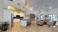 Located at 1055 W Baseline Rd in Mesa, Arizona, this single-floor open-concept community space combines a fully equipped kitchen with a generously sized living area, all unified by warm wood-look tile flooring throughout. The interior is immediately distinguished by its soaring vaulted ceilings, which lend an airy, expansive feel and are complemented by an abundance of recessed lighting and ceiling fans for year-round comfort.

The kitchen is outfitted with crisp white shaker cabinetry, dark countertops, a dark tile backsplash, stainless steel appliances including a full-size refrigerator, and a central island with bar seating — ideal for casual gatherings. Vibrant artwork adds a pop of color to the walls, reinforcing the welcoming atmosphere.

The living area is open and versatile, furnished with a mix of neutral and teal-accented seating arrangements centered around a natural wood coffee table. A wall-mounted television, a decorative display cabinet, and carefully placed greenery contribute to the room's inviting character. A distinctive arched doorway frames a view into an adjacent glass-enclosed area.

Large glass doors and floor-to-ceiling windows along the rear wall flood the space with natural light and offer direct views of an outdoor pool and patio area, seamlessly connecting the interior to the outdoors.