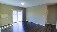 This single-floor space presents a clean, functional layout with a straightforward design aesthetic. The main living area is finished with rich, dark hardwood flooring that contrasts neatly against the neutral beige-toned walls, creating a grounded, cohesive look throughout. White baseboard trim lines the perimeter of the room, adding a crisp finishing detail.

A prominent sliding glass door serves as a key architectural feature, allowing natural light to pour into the space and offering direct views to the exterior. The door is framed with white trim and fitted with a sheer curtain panel, softening the incoming light while maintaining an open, airy feel.

Baseboard heating runs along the exterior wall beneath the sliding door, providing a practical heating solution integrated seamlessly into the room's layout. A flush-mount ceiling light fixture provides overhead illumination, and a wall-mounted control panel is visible near the entry area, suggesting climate or electrical management.

The walls are smooth and painted in a warm greige tone, offering a versatile backdrop suitable for a variety of uses. Overall, the space feels open, well-lit, and move-in ready, with durable finishes well-suited for everyday living.