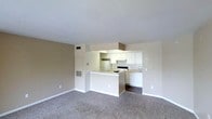 This single-floor space offers an open-concept layout that seamlessly connects the living area and kitchen, creating a cohesive and functional environment. The living area is finished with wall-to-wall carpeting in a neutral tone, providing a comfortable foundation that complements the light-colored walls throughout.

The kitchen is thoughtfully designed with white cabinetry offering ample storage, a white electric range, and a full-size refrigerator. A peninsula breakfast bar serves as a natural divider between the kitchen and living space, adding both a casual dining option and additional counter space. The kitchen flooring transitions to a darker hard surface, distinguishing it from the carpeted living area.

Overhead, a built-in range hood is positioned above the cooking area, while recessed ventilation registers are visible in both the ceiling and walls, indicating a central HVAC system throughout the unit. The smooth white ceilings and clean wall finishes contribute to a bright and airy atmosphere. The overall design is clean and practical, with a neutral color palette that lends itself well to a variety of furnishing styles. The space is well-suited for everyday living, with an efficient flow between its core functional areas.