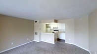 This single-floor space offers an open-concept layout that seamlessly connects the living area and kitchen, creating a cohesive and functional environment. The living area is finished with wall-to-wall carpeting in a neutral tone, providing a comfortable foundation that complements the light-colored walls throughout.

The kitchen is thoughtfully designed with white cabinetry offering ample storage, a white electric range, and a full-size refrigerator. A peninsula breakfast bar serves as a natural divider between the kitchen and living space, adding both a casual dining option and additional counter space. The kitchen flooring transitions to a darker hard surface, distinguishing it from the carpeted living area.

Overhead, a built-in range hood is positioned above the cooking area, while recessed ventilation registers are visible in both the ceiling and walls, indicating a central HVAC system throughout the unit. The smooth white ceilings and clean wall finishes contribute to a bright and airy atmosphere. The overall design is clean and practical, with a neutral color palette that lends itself well to a variety of furnishing styles. The space is well-suited for everyday living, with an efficient flow between its core functional areas.