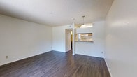 This single-floor space features an open-concept layout that seamlessly connects the living area to the kitchen. The living area is finished with dark hardwood-style flooring that contrasts cleanly against the white walls, creating a bright and modern aesthetic. A textured popcorn ceiling adds a classic touch overhead.

The kitchen is positioned at the far end of the space and is accessible via a pass-through breakfast bar, which also serves as a visual divider between the two areas. A pendant light hangs above the bar, providing focused illumination and a subtle design element. The kitchen itself includes visible appliances and cabinetry, offering functional storage and cooking space.

A doorway leads to an adjacent hallway or room, suggesting additional private spaces beyond the main living area. The open layout allows natural light to filter through, contributing to an airy and welcoming ambiance. The clean, neutral color palette throughout provides a versatile backdrop suitable for a variety of interior styles. Overall, the space balances practicality with a straightforward, modern design.