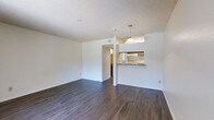 This single-floor space features an open-concept layout that seamlessly connects the living area to the kitchen. The living area is finished with dark hardwood-style flooring that contrasts cleanly against the white walls, creating a bright and modern aesthetic. A textured popcorn ceiling adds a classic touch overhead.

The kitchen is positioned at the far end of the space and is accessible via a pass-through breakfast bar, which also serves as a visual divider between the two areas. A pendant light hangs above the bar, providing focused illumination and a subtle design element. The kitchen itself includes visible appliances and cabinetry, offering functional storage and cooking space.

A doorway leads to an adjacent hallway or room, suggesting additional private spaces beyond the main living area. The open layout allows natural light to filter through, contributing to an airy and welcoming ambiance. The clean, neutral color palette throughout provides a versatile backdrop suitable for a variety of interior styles. Overall, the space balances practicality with a straightforward, modern design.