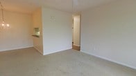This single-floor residence presents a straightforward, functional layout designed to maximize usable living space. The main living area is finished with wall-to-wall carpet in a neutral tone, contributing to a quiet and comfortable atmosphere. Light-colored walls throughout lend the space a clean, airy feel, helping to reflect ambient light and enhance the sense of openness.

A partial dividing wall separates the living area from the kitchen, which features a dark-topped breakfast bar counter that provides casual dining or workspace. The kitchen area, visible from the main living space, includes cabinetry with a practical, understated design. A ceiling fan is installed overhead in the living area, supporting year-round climate comfort.

A doorway opening on one side leads to additional rooms, suggesting a connected layout that flows naturally between living, kitchen, and private spaces. White baseboards frame the carpeted floors throughout, adding a crisp finishing detail. The overall ambiance is modest and move-in ready, with a neutral palette that serves as a versatile backdrop for a variety of interior styles.