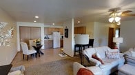 This single-floor space offers a thoughtfully designed open-concept layout that integrates the kitchen, dining, and living areas into one cohesive environment. The kitchen is outfitted with warm wood cabinetry, a white refrigerator, and a double sink, complemented by recessed lighting that keeps the space bright and functional. A small appliance station with a coffee maker adds everyday convenience. Adjacent to the kitchen, a raised bar-height dining area with dark bar stools provides a casual spot for meals or entertaining. A separate dining nook features a glass-top table with matching dark chairs, accented by a fresh floral centerpiece that adds a welcoming touch. The living area is anchored by plush white leather seating with rust-toned accent pillows, creating a comfortable and inviting atmosphere. A large area rug defines the lounge space, adding warmth to the flooring underfoot. A central support column subtly divides the space while maintaining the open, airy feel throughout. Recessed lighting is consistent across the entire floor, ensuring even illumination throughout. The neutral color palette of whites, beiges, and warm wood tones contributes to a clean, cohesive aesthetic that feels both practical and inviting.