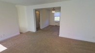 Located at 1 Martha Avenue in Wrightstown, NJ, this single-floor residence offers a straightforward and functional layout with a clean, neutral aesthetic throughout. The interior features wall-to-wall carpeting in a warm beige tone that extends seamlessly across the open living areas, providing a comfortable and cohesive feel underfoot. The walls are finished in crisp white, enhancing the sense of brightness and openness throughout the space. A ceiling fan with a light fixture is centrally positioned, offering both ventilation and ambient lighting to the main living area. Natural light filters in through a window framed by a window treatment, adding a connection to the outdoors. A radiator unit beneath the window indicates a reliable heating system built into the home. The open flow between rooms creates an easy sense of movement through the space, with a hallway visible in the background leading to additional areas of the home. The overall ambiance is clean, neutral, and move-in ready, providing a solid foundation for personalization. The single-level layout offers practical, accessible living in a quiet residential setting in Wrightstown, NJ.