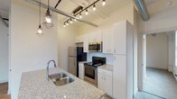 Unit 1102 is a thoughtfully designed urban loft that masterfully combines industrial heritage with contemporary living. The open layout is anchored by warm hardwood flooring that flows through the main living areas, while exposed white-painted brick walls and prominent silver ductwork running along the ceilings lend an authentic loft aesthetic throughout.

The kitchen is a standout feature, outfitted with white shaker cabinetry, granite countertops, a subway tile backsplash, and a full suite of stainless steel appliances including a refrigerator, electric range, and built-in microwave. An oversized granite-topped island with a double stainless steel sink serves as both a functional workspace and a natural gathering point. Industrial-style cage pendant lighting adds character overhead.

The generously sized living area opens to corner windows that frame impressive city skyline views, filling the space with natural light. The bedroom features plush carpeting, whitewashed brick walls, and a window with additional urban views, creating a calm, comfortable retreat. The bathroom is clean and well-appointed with a granite vanity, a full tub-shower combination, and light wood-look flooring. Exposed ductwork and high ceilings throughout reinforce the loft's distinctive industrial charm.