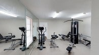 Located at 1651 Knollwood Dr in Mobile, Alabama, this fitness center offers a practical and well-equipped space for exercise and physical wellness. The room is finished with wall-to-wall carpeting that provides a comfortable surface underfoot, while a textured ceiling and bright fluorescent lighting ensure the space is clearly illuminated throughout. A full-length mirrored wall runs along one side of the room, visually expanding the space and allowing users to monitor their form during workouts. Natural light supplements the overhead lighting through a window fitted with horizontal blinds. The facility is outfitted with a variety of exercise equipment, including a treadmill, an elliptical cross-trainer, a multi-station cable and weight machine, an adjustable workout bench, and additional free weight equipment. An emergency exit door is positioned at the rear of the room, contributing to the safety and functionality of the space. The neutral color palette of beige walls and light carpeting creates a clean, distraction-free environment conducive to focused exercise. Overall, this fitness center provides a compact yet well-appointed space that addresses a broad range of cardiovascular and strength training needs.
