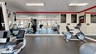 Located at 213 Lingstorm Ln in North Chesterfield, VA, this fitness center offers a clean, well-organized single-floor layout designed to support a full range of workout activities. The space features a bright, open exercise area filled with an array of equipment, including treadmills, stationary bikes, a multi-station strength machine, an elliptical, and free weights, providing options for both cardio and resistance training. Large floor-to-ceiling mirrors run along the interior walls, visually expanding the space and allowing users to monitor their form during workouts. A bold red accent stripe runs along the upper perimeter of the walls, adding a dynamic contrast to the neutral beige tones. The flooring throughout is a durable, dark rubber surface suited for heavy equipment and high-traffic use. A series of large windows lines the exterior wall, and a glass entry door floods the interior with abundant natural light while offering views of the surrounding greenery and outdoor environment. Flush-mounted ceiling light panels complement the natural light, ensuring consistent illumination throughout. A textured ceiling and beadboard wainscoting detail along the lower walls contribute a structured, clean aesthetic to the overall design of this functional and inviting fitness facility.