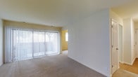This single-floor residence features a spacious, open living area that serves as the central focus of the space. The room is finished with neutral-toned wall-to-wall carpeting, contributing to a clean and understated aesthetic. A large sliding glass door spans a significant portion of one wall and is dressed with vertical blinds, allowing for adjustable natural light and providing direct access to the outdoors. The abundant glazing ensures the living area is well-lit throughout the day, creating a bright and airy atmosphere. The walls are painted in crisp white and soft warm tones, lending the space a light, open feel. An adjoining room, visible through an open doorway, features warm yellow-toned walls and an additional window, further enhancing the natural light within the unit. The layout appears open and functional, with a clear sightline between the main living area and the adjacent space. Clean, simple architectural lines and neutral finishes throughout provide a versatile backdrop suitable for a variety of interior styles. The overall ambiance is calm, uncluttered, and move-in ready.