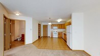 This single-floor space offers a functional and open layout, seamlessly connecting the kitchen and living areas. The kitchen is well-appointed with warm honey-toned wood cabinetry providing ample storage, light-colored countertops, and a full suite of white appliances including a refrigerator, electric range, and built-in microwave. A pendant light fixture hangs above, adding a focal point to the kitchen area. The flooring transitions from durable ceramic tile in the kitchen to plush carpeting in the adjacent living space, clearly delineating the two zones while maintaining an open feel. The textured white ceiling and neutral white walls throughout contribute to a bright and clean atmosphere. A doorway visible from the main area leads to additional spaces, including what appears to be a walk-in closet or storage area, suggesting practical storage solutions throughout. The open-concept design allows for flexible furniture arrangement and promotes a sense of spaciousness, making the space feel inviting and easy to navigate.