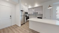 This single-floor unit showcases a clean, contemporary design with a cohesive neutral palette and durable light-toned luxury vinyl plank flooring running throughout the main living areas. The open-concept layout connects the living space directly to the kitchen, which serves as a functional centerpiece of the unit. The kitchen is equipped with stainless steel appliances, including a refrigerator, gas range, microwave, and dishwasher. White flat-front cabinetry extends floor to ceiling, providing ample storage, while a light grey subway tile backsplash adds subtle texture. A central island with a white quartz countertop offers additional prep space and casual seating potential. Pendant lighting and recessed fixtures illuminate the area effectively. Large windows in the living area allow natural light to fill the space, offering views of the surrounding neighborhood. The bedroom features plush grey carpeting and a sliding glass door that opens to an outdoor patio, enhancing connectivity to the exterior. The full bathroom is well-appointed with a wide white vanity, quartz countertop, large mirror, and a combination shower and soaking tub. The overall ambiance is bright, clean, and move-in ready, reflecting a modern residential aesthetic.