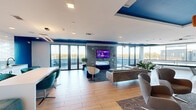 Located at 3061 George Busbee Pkwy NW in Kennesaw, GA, this contemporary community lounge is a well-appointed, single-floor space designed for socializing and recreation. The open-plan layout is anchored by a generous living area furnished with modern sofas, accent chairs in teal and grey, and a bold blue ottoman, all centered around a sculptural hexagonal-tile feature wall with a wall-mounted flat-screen TV and a linear fireplace below. Large floor-to-ceiling sliding glass doors flood the space with natural light and open directly onto an outdoor terrace, offering sweeping panoramic views of the surrounding area. The flooring throughout is light wood-tone tile, lending warmth to the contemporary aesthetic. A dedicated game zone features a full-size Kettler ping-pong table beneath a sleek linear pendant light, with additional wall-mounted screens for entertainment. A built-in workstation area with a long white desk, teal task chairs, and cabinetry provides a functional co-working nook. Bold cobalt blue wall accents and ceiling details tie the color palette together throughout, creating a cohesive and energetic atmosphere. Recessed lighting and multiple mounted TVs ensure the space is equally functional day and night.