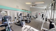This fitness center offers a well-organized and functional space designed to support a variety of workout routines. The layout provides ample room to move between equipment comfortably, creating an open and accessible environment for exercise. Natural light streams in through multiple windows fitted with shutters, brightening the space and contributing to an energizing atmosphere. A ceiling fan overhead aids in air circulation, keeping the environment comfortable during workouts. The flooring features durable, rubberized gym matting, providing both safety and stability underfoot. A prominent mirrored wall with a teal accent stripe runs along one side of the room, visually expanding the space while allowing users to monitor their form. The equipment selection includes elliptical trainers, a cable machine, a multi-function strength training station, and additional cardio machines, offering a comprehensive range of exercise options. The walls are painted in a clean, neutral tone, lending the space a fresh and modern feel. Recessed fluorescent lighting supplements the natural light, ensuring the room remains well-lit at all times. Overall, this fitness center is a practical and inviting space, thoughtfully designed to accommodate both cardio and strength training needs in a single, streamlined environment.