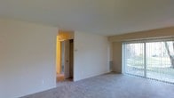 Unit 102 is a single-floor residence that offers a clean, functional layout designed for comfortable everyday living. The main living area is spacious and open, finished with soft wall-to-wall carpeting that adds warmth underfoot. A large sliding glass door fitted with vertical blinds allows an abundance of natural light to pour into the space while providing a pleasant view of the surrounding greenery outside. The neutral, light-colored walls create a bright and airy ambiance throughout, offering a versatile canvas for personalization. A hallway connects the living area to the rest of the unit, leading to additional rooms and storage spaces. The inclusion of closet space visible through the hallway indicates thoughtful storage solutions within the layout. The overall design is straightforward and practical, with a consistent aesthetic maintained across the space. The single-floor layout ensures easy accessibility, making movement between areas seamless. The combination of natural light, open floor space, and neutral finishes gives this unit a welcoming and move-in ready feel.