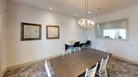 This well-appointed single-floor space functions as a versatile meeting and gathering area, combining refined design with practical functionality. The main room is anchored by a substantial dark wood conference-style table surrounded by white ladder-back chairs, creating a welcoming yet professional atmosphere. A striking gold ring chandelier with glass cylinder accents serves as the room's focal point, casting warm, even light throughout the space.

Along one wall, a sleek built-in marble-topped desk provides additional workspace, accompanied by modern black task chairs — ideal for individual work or small group collaboration. Patterned carpet in soft blue and grey tones adds texture and visual interest underfoot, complementing the neutral white walls. Natural light filters in through a curtained window dressed in geometric-patterned drapes, enhancing the bright, open feel of the room.

Curated wall art, including framed map prints and bold abstract paintings in deep blues and warm ambers, lends the space a creative and cultured character. A large framed mirror on one wall amplifies the sense of openness. An adjoining room, accessible through a glass-paneled grey door, features decorative diamond-patterned tile and a vanity area, rounding out this thoughtfully designed, multi-functional space.