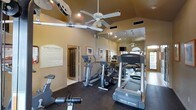 This fitness center is designed to support a well-rounded exercise routine within a compact, efficiently arranged space. The room features vaulted ceilings that lend an open, airy feel, complemented by a ceiling fan that helps maintain comfortable airflow during workouts. Recessed lighting provides consistent illumination throughout.

The floor is finished in a dark, durable material suited for high-traffic exercise use. A full-length mirrored wall lines one side of the room, visually expanding the space and allowing users to monitor their form during workouts. The cardio equipment selection includes treadmills, an elliptical trainer, a stair climber, and a recumbent bike, offering a variety of aerobic training options.

Warm, neutral-toned walls contribute to an inviting atmosphere. A posted Fitness Center Rules sign near the entrance helps establish guidelines for shared use of the facility. Framed artwork and a window with a door providing natural light add to the welcoming environment. The overall design balances functionality with comfort, making it a practical and pleasant space for daily exercise.