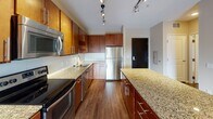 Located at 425 Main St SE in Minneapolis, Minnesota, this well-appointed single-floor unit offers a thoughtfully designed open-concept layout that seamlessly connects the kitchen and living area. The kitchen is a standout feature, showcasing rich warm-toned cabinetry, granite countertops, a full suite of stainless steel appliances including a refrigerator, range, microwave, and dishwasher, and a large central island with additional counter space. Track lighting illuminates the kitchen and living areas, complementing the warm hardwood flooring that runs throughout the main living space. The living area is generously sized and bathed in natural light from a floor-to-ceiling sliding glass door that opens to a private balcony with outdoor views of the surrounding Minneapolis neighborhood and greenery. A carpeted bedroom accessed from the main living space provides a comfortable retreat. The bathroom features matching granite countertops, a dual-sink vanity with warm wood cabinetry, and a large mirror. Neutral wall tones and textured ceilings complete the interior aesthetic, creating a cohesive and modern atmosphere throughout this urban Minneapolis residence.