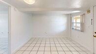 Located in Miami, FL, this single-floor, one-bedroom, one-bathroom unit offers a clean and functional layout finished consistently with large white ceramic tiles throughout. The kitchen is efficiently designed, featuring white cabinetry extending to the ceiling for ample storage, a warm earth-toned mosaic tile backsplash, a white electric range, a refrigerator, and a single-basin sink. The open layout allows a natural flow from the kitchen into the living and sleeping areas. The bedroom is generously sized relative to the overall footprint, offering a bright, open feel with a window that brings in natural light and white walls that enhance the airy atmosphere. A dedicated closet provides practical storage. The bathroom includes a granite-topped vanity, a tiled walk-in shower, and a window for natural ventilation and light. The shower wall tiles feature a distinctive accent pattern with colorful square tile details in red, blue, and green, adding a retro character to the space. Throughout the unit, the consistent white palette and tile flooring create a cohesive, easy-to-maintain environment well-suited to Miami's warm climate.