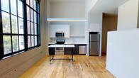 This single-floor space showcases a compelling blend of industrial heritage and contemporary design. The open-concept layout is anchored by soaring ceilings that amplify the sense of space, while original exposed concrete walls and large black-framed industrial windows flood the interior with natural light and offer views of surrounding greenery. Warm hardwood flooring runs throughout the main living areas, complementing the raw architectural elements.

The kitchen is well-appointed with stainless steel appliances, including a range, microwave, refrigerator, and dishwasher. Dark lower cabinetry is paired with white upper cabinets and a light stone countertop, creating a modern two-tone aesthetic. A suspended linear pendant light above a mobile island adds functional workspace and a sleek, contemporary touch.

A half-wall partition defines the sleeping area, maintaining an open flow while providing a degree of separation. The bathroom features dark wood-look tile flooring, a dark vanity with a white countertop, an undermount sink, and a backlit mirror, delivering a clean, modern feel.

Additional highlights include an in-unit stacked washer and dryer, a dedicated closet with wire shelving, and a ceiling fan to enhance comfort throughout the space.