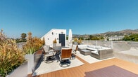 Located at 1257 N Detroit St in West Hollywood, CA, this rooftop deck offers an exceptional outdoor living experience with sweeping panoramic views of the Hollywood Hills and the surrounding Los Angeles cityscape. The deck features warm-toned composite wood flooring that extends across the entire space, creating a cohesive and inviting atmosphere under the abundant Southern California sunshine.

The rooftop is thoughtfully organized into two distinct functional zones. A generous lounge area is anchored by a large L-shaped wicker sectional sofa with plush neutral cushions, paired with a matching coffee table — ideal for relaxed gatherings. Adjacent to it, a dedicated outdoor dining area accommodates a full-sized rectangular dining table surrounded by cushioned swivel chairs with woven leather accents, complemented by a gas grill nearby for al fresco entertaining.

Tall planters filled with ornamental grasses and drought-tolerant greenery line the perimeter, adding texture and a sense of privacy. A glass and mesh railing system runs along the edges, preserving unobstructed sightlines to the hills and city beyond. The open-sky exposure ensures the space is bathed in natural light throughout the day, making it a compelling venue for both casual relaxation and elevated outdoor entertaining in the heart of West Hollywood.