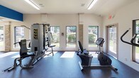 Located at Villas of Hulen Bend in Fort Worth, Texas, this single-floor fitness center is designed to support a well-rounded workout experience. The space is divided into two connected zones, each equipped with a distinct category of exercise equipment, allowing users to move seamlessly between cardio and strength training.

The main workout area features rubberized flooring throughout, providing both safety and durability underfoot. Cardio equipment includes a True treadmill and a True upright stationary bike, positioned to take advantage of the natural light streaming through multiple large windows that overlook the surrounding greenery. A multi-station cable machine offers a range of resistance training options, complemented by a dedicated free weight area housing a full rack of dumbbells and an adjustable workout bench.

The walls are painted white with bold blue accent graphics and motivational words — "FIT," "STRENGTH," and "FOCUS" — lending the space an energetic and purposeful atmosphere. Overhead fluorescent lighting ensures consistent illumination throughout. A secondary open area adjoins the main gym, bright with floor-to-ceiling windows, providing additional functional space. Fitness center rules are posted near the entrance, and the layout is efficient and easy to navigate, making this facility a practical and well-appointed amenity.