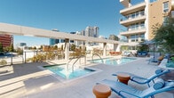 This outdoor amenity deck is set within a contemporary multi-story residential building and offers a well-appointed retreat above the city. The centerpiece of the space is a generously sized swimming pool with crystal-clear blue water, complemented by a separate spa/hot tub, both finished with clean, modern tiling and equipped with stainless steel entry rails. The surrounding deck is paved in light-toned concrete, providing ample space for lounging with an array of blue chaise lounges and terracotta-hued side tables arranged throughout. Shade structures, including a striped pergola and market umbrellas, offer relief from the sun, while additional seating areas with outdoor sofas and dining sets make the deck suitable for both relaxation and social gatherings. Lush planters with ornamental trees and shrubs add a natural touch to the hardscape environment. Glass-panel railings line the perimeter, ensuring unobstructed panoramic views of the surrounding urban skyline. The building's modern facade, featuring balconies with glass railings and large windows, frames the deck on multiple sides. The overall ambiance is bright, open, and sophisticated, combining resort-style comfort with an urban setting.