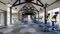 This single-story fitness center is defined by its striking architectural design, highlighted by a dramatic vaulted ceiling with exposed dark wood beam trusses that create an open, airy atmosphere. Pendant lighting hangs from the ceiling structure, providing warm, evenly distributed illumination throughout the space. The expansive floor plan accommodates a wide variety of exercise equipment, including a row of treadmills, elliptical machines, rowing machines, and strength-training equipment, all arranged to allow comfortable movement between stations. One of the most distinctive design elements is the eye-catching hexagonal tile flooring in a blend of green, gray, and blue tones, adding a modern, geometric aesthetic to the interior. Large windows along one wall flood the space with natural light and offer views of the surrounding landscape. A glass-paneled door at the far end of the room provides additional access and reinforces the open, welcoming feel of the space. The combination of rustic wood elements, contemporary finishes, and abundant natural light creates a motivating and visually engaging environment well-suited for fitness and wellness activities.