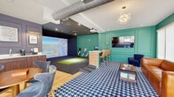 Located at 3000 Strand Way in Lancaster, PA, this single-floor entertainment lounge is thoughtfully designed to blend leisure and recreation in a compact yet well-utilized space. The centerpiece is a full indoor golf simulator bay, complete with a large projection screen, a dedicated hitting mat, and a wood-paneled control counter with bar-height seating — ideal for players and spectators alike. A ceiling-mounted projector delivers an immersive golfing experience, while track lighting illuminates the simulator zone against its dark, contrast-rich backdrop. The lounge area is anchored by a rich cognac leather sofa, navy accent chairs, and a bold blue-and-white houndstooth area rug over warm hardwood flooring, lending the space a lively, contemporary character. Vibrant teal wainscoting and paneled walls define the lounge and entryway, accented by a decorative bubble chandelier and wall-mounted flat-screen television. A dedicated wet bar features rich wood cabinetry, a sink, countertop appliances, and framed golf-themed artwork displayed under brass picture lights. The entry area offers large windows with roller shades, allowing natural light to filter in. Exposed ductwork on the ceiling adds an industrial touch that complements the space's eclectic, upscale aesthetic.