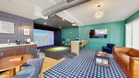 Located at 3000 Strand Way in Lancaster, PA, this single-floor entertainment lounge is thoughtfully designed to blend leisure and recreation in a compact yet well-utilized space. The centerpiece is a full indoor golf simulator bay, complete with a large projection screen, a dedicated hitting mat, and a wood-paneled control counter with bar-height seating — ideal for players and spectators alike. A ceiling-mounted projector delivers an immersive golfing experience, while track lighting illuminates the simulator zone against its dark, contrast-rich backdrop. The lounge area is anchored by a rich cognac leather sofa, navy accent chairs, and a bold blue-and-white houndstooth area rug over warm hardwood flooring, lending the space a lively, contemporary character. Vibrant teal wainscoting and paneled walls define the lounge and entryway, accented by a decorative bubble chandelier and wall-mounted flat-screen television. A dedicated wet bar features rich wood cabinetry, a sink, countertop appliances, and framed golf-themed artwork displayed under brass picture lights. The entry area offers large windows with roller shades, allowing natural light to filter in. Exposed ductwork on the ceiling adds an industrial touch that complements the space's eclectic, upscale aesthetic.
