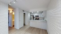 This two-floor space showcases a clean, contemporary design with a neutral color palette and light hardwood flooring that runs throughout the main level. The kitchen is a central highlight, featuring white shaker-style cabinetry, light granite countertops, and a full suite of stainless steel appliances, including a refrigerator, microwave, and gas range. A peninsula with vertical shiplap paneling extends from the kitchen, providing additional counter space and serving as a natural divider between the kitchen and the adjacent living area. Recessed lighting above the kitchen ensures the workspace is well-lit, complementing the natural brightness of the overall space. A textured ceiling adds subtle dimension overhead. Adjacent to the kitchen, an open doorway leads to a built-in shelving unit, offering practical storage solutions. A louvered closet door nearby provides additional storage access. The open layout promotes a sense of flow between functional areas, while the consistent light tones of the walls, cabinetry, and flooring contribute to a bright and airy ambiance throughout the main level of this two-floor space.