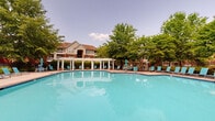 Located at 1200 Virginia Center Pkwy in Glen Allen, Virginia, this single-level amenity space offers residents a well-appointed fitness center and a resort-style outdoor swimming pool. The fitness center is designed with a vaulted ceiling that enhances the sense of openness, while a bold teal accent wall adds a vibrant, energizing atmosphere. Wood-look flooring runs throughout the space, providing a clean and modern aesthetic. Natural light flows in through multiple windows fitted with white blinds, including a distinctive arched window that serves as a focal point. The facility is equipped with a comprehensive range of exercise equipment, including treadmills, elliptical machines, a multi-functional cable machine, a Spirit treadmill, medicine balls, and a dumbbell rack, making it suitable for a variety of workout routines. A glass-paneled door provides direct access to the outdoor area. Outside, the expansive swimming pool features a freeform shape with clear turquoise water, surrounded by a generous pool deck furnished with lounge chairs in a matching teal color. A pergola structure with columns offers shaded seating, while lush mature trees frame the area, creating a private and serene outdoor retreat. Together, these amenities provide a comprehensive and inviting environment for recreation and wellness.