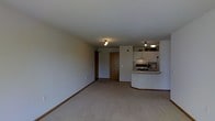 This single-floor space offers a clean and functional layout designed for comfortable everyday living. The main living area is finished with wall-to-wall carpeting and neutral-toned walls, creating a bright and understated ambiance throughout. Ceiling-mounted light fixtures provide consistent illumination across the open floor plan.

The kitchen is positioned toward the rear of the main living space and features white upper cabinetry with open shelving above, a range with an overhead hood, and a curved breakfast bar that serves as both a workspace and a casual dining surface. The pass-through design of the kitchen keeps the space feeling open and connected to the living area.

A wood-toned entry door anchors the back wall, offering a clearly defined entryway that transitions naturally into the living space. The layout flows efficiently, making good use of available square footage without feeling cramped. White baseboards line the perimeter of the room, adding a clean finishing detail throughout.

Overall, the space presents a straightforward, well-maintained interior with a practical open-concept design that balances the kitchen and living areas cohesively.