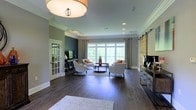 This elegant single-floor space showcases a thoughtfully designed interior that balances classic architectural detail with contemporary style. The main living area is anchored by rich, dark hardwood flooring that flows throughout, lending warmth and continuity to the space. Crown molding lines the ceilings, adding a refined architectural touch that complements the neutral gray wall palette, accented by a striking deep teal feature wall.

Natural light pours in through a large bank of windows dressed with patterned drapery panels, illuminating the open layout and creating a bright, welcoming atmosphere. A set of white upholstered armchairs arranged around a round coffee table forms an inviting seating vignette, ideal for conversation and relaxation. A sliding barn door adds a rustic yet stylish contrast to the otherwise polished interior.

Recessed lighting and a bold drum-shade flush mount fixture provide layered illumination, enhancing the space's ambiance at any time of day. A built-in media console and a French door leading to an adjacent area further highlight the functional versatility of the layout. Orange accent pillows and greenery provide vibrant pops of color, completing the sophisticated and inviting character of this well-appointed space.