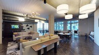 This dynamic game room offers a thoughtfully designed open-concept space that seamlessly blends relaxation and social gathering. The room is anchored by a generous lounge area furnished with plush, neutral-toned sofas and accent pillows in warm mustard hues, arranged around a rustic wood coffee table that adds an organic contrast to the contemporary setting. Adjacent to the lounge, a dining and gathering zone features round tables paired with sleek, modern chairs, ideal for group activities or casual conversation. The space is distinguished by its dramatically textured geometric ceiling, which adds a bold architectural statement overhead. A mix of pendant lighting fixtures — including large cylindrical drum shades and a sculptural brass chandelier — create layered, ambient illumination throughout. Dark hardwood-style flooring grounds the space with warmth and depth. Floor-to-ceiling windows line the perimeter, flooding the interior with abundant natural light and offering views of the surrounding cityscape. Abstract wall art adds an artistic touch, enhancing the modern industrial aesthetic. The result is a versatile, well-appointed space that encourages both lively entertainment and relaxed social interaction.