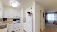 This two-floor residential space offers a thoughtfully arranged layout designed for comfortable everyday living. The kitchen is a standout feature, showcasing white cabinetry that extends to the upper walls, providing ample storage space. A full-size white refrigerator and electric range with coil burners are seamlessly integrated into the kitchen design, complemented by light-toned countertops. A structural partition wall adds visual definition to the kitchen, creating a sense of organization and flow between spaces. The flooring is light and neutral, contributing to the overall bright and airy atmosphere. Flush-mounted ceiling lighting ensures the kitchen is well-lit, while a ceiling vent indicates a central HVAC system for year-round climate control. The color palette throughout is predominantly white and off-white, lending a clean, open feel to the interiors. A hallway visible from the kitchen leads further into the home, hinting at additional rooms and living areas across the two floors. The overall ambiance is practical and welcoming, with a neutral aesthetic that provides a versatile backdrop for a variety of interior styles.