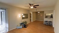 This single-floor space offers an open, connected layout that flows naturally between the living area, dining space, and kitchen. The living room is anchored by warm hardwood-style flooring that extends throughout, complemented by neutral taupe walls that create a calm, cohesive atmosphere. A ceiling fan with an integrated light fixture is centrally mounted overhead, while a textured ceiling adds subtle architectural character.

The open layout allows the living area to transition seamlessly into a dining space, which features a pendant light fixture and provides direct sightlines into the adjacent kitchen. The kitchen, partially visible from the main living area, includes cabinetry and counter seating, suggesting a practical and accessible design.

A doorway from the living room leads to an additional area — likely a hallway connecting to further rooms — adding depth and versatility to the floor plan. Decorative wall art and accent lighting add warmth to the space. Consistent flooring, neutral tones, and an open configuration make this a functional and inviting single-level home.