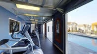 This single-floor fitness center offers a thoughtfully designed workout space that seamlessly blends indoor and outdoor living. The standout architectural feature is the large retractable garage-style door that opens fully to the adjacent outdoor pool area, flooding the space with natural light and fresh air. The interior is finished with deep navy blue walls and dark rubber flooring, creating a sleek, focused atmosphere ideal for exercise. A row of cardio machines, including ellipticals and treadmills, lines one side of the room, offering ample space for a full workout routine. Strength and multi-use training equipment is visible toward the rear of the space, rounding out the facility's offerings. An ornate wood-framed mirror mounted on the wall adds a distinctive decorative touch against the bold color palette. Overhead, exposed industrial-style ceiling tracks and fluorescent lighting contribute to the space's modern, utilitarian aesthetic. When the garage door is raised, the gym opens directly onto a sunlit pool deck enclosed by wrought iron fencing, with views of the surrounding community beyond — creating a uniquely connected indoor-outdoor fitness experience.