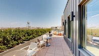 This rooftop terrace offers a compelling blend of contemporary design and open-air functionality. The space is defined by a striking metal-clad structure with floor-to-ceiling glass curtain walls, allowing natural light to flood adjacent interior areas while providing transparency between indoors and out. The terrace wraps around the structure, offering generous tiled outdoor areas suited for a variety of uses.

Multiple distinct seating zones are distributed across the terrace, furnished with a mix of colorful Adirondack chairs, modern slatted benches, and bistro-style table-and-chair sets, creating informal gathering nodes throughout the space. Perforated metal railings and low planter walls lined with lush hedging define the perimeter, contributing both safety and a touch of greenery against the urban backdrop.

The panoramic city views are a standout feature, with a historic water tower, a prominent brick chimney stack, downtown skyline buildings, and open sky all visible from various vantage points. Wall-mounted exterior lighting and security fixtures are integrated into the façade, underscoring the space's practical design. The open layout, combined with abundant natural light and sweeping urban vistas, creates an engaging and versatile outdoor environment well-suited for communal use.