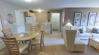 This single-floor unit features an open-concept layout that seamlessly connects the kitchen, dining area, and living room into one cohesive space. The kitchen is equipped with light wood cabinetry, a white refrigerator, and a gas range, with a peninsula counter that serves as a natural divider between the kitchen and the rest of the space. Adjacent to the kitchen, a cozy dining nook accommodates a round wooden table with cushioned chairs, ideal for casual meals. The living area is carpeted and finished in warm taupe and beige tones, creating a welcoming ambiance. A plush upholstered sofa and armchair are positioned to encourage comfortable conversation, accompanied by a floor lamp that adds soft ambient lighting. A framed wall piece adds a decorative accent to the living room wall. A hallway visible in the background leads to additional areas of the unit, hinting at further functional spaces beyond. The overall design emphasizes a practical and comfortable layout, with consistent neutral finishes throughout that contribute to a calm and understated aesthetic.