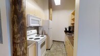 This 1-bedroom, 1-bathroom residence is thoughtfully arranged on a single floor, making efficient use of its layout. The galley-style kitchen is a standout feature, designed to maximize functionality within a compact footprint. It is outfitted with a full suite of white appliances, including an electric coil range, over-the-range microwave, and a full-size refrigerator. Light wood cabinetry lines the upper and lower walls, providing ample storage, while open shelving adds accessible display space. A stainless steel sink is positioned along the counter on the opposite wall, complemented by a dark-toned countertop that offers a subtle contrast to the light cabinetry. The flooring throughout the kitchen consists of neutral tile, contributing to a clean and practical aesthetic. Exposed structural elements along the walls add a touch of character to the space. Overhead fluorescent lighting ensures the kitchen remains bright and well-lit. The overall design of this unit prioritizes practicality and ease of use, making it a well-suited option for those seeking a straightforward and efficiently organized living space.