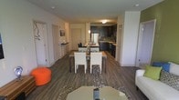 This well-appointed single-floor space showcases a thoughtfully designed open-concept layout that seamlessly connects the main living, dining, and kitchen areas. Rich, dark hardwood-style flooring runs throughout, grounding the space with a warm yet contemporary feel.

The dining area takes center stage, featuring a rectangular table paired with crisp white upholstered chairs and dark metal legs, creating a clean contrast. A pendant light fixture hangs above, adding a refined touch to the space. Adjacent to the dining area, a small breakfast bar with bar stools offers an additional casual seating option.

The kitchen is outfitted with dark cabinetry that extends to the upper and lower cabinets, stainless steel appliances, and light countertops that provide a striking contrast against the darker tones. The layout offers ample counter space and functional storage throughout.

The living area, visible in the foreground, features a neutral-toned sofa and a low-profile coffee table set atop a textured area rug, while a bold orange accent ottoman adds a pop of color and personality. Bright white walls and recessed lighting keep the interior feeling open and airy. Multiple interior doors suggest well-distributed private rooms beyond the central living space.
