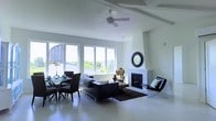 This contemporary single-floor space showcases a clean, modern aesthetic defined by a crisp white palette and polished concrete flooring that flows seamlessly throughout. The open-plan layout integrates the living and dining areas into one cohesive, light-filled environment. Large windows along the exterior wall flood the space with natural light while framing views of the surrounding greenery and neighborhood beyond, creating a strong visual connection to the outdoors. The living area is anchored by a sleek marble fireplace with a classic circular mirror mounted above, serving as an elegant focal point. A ceiling fan overhead adds both functionality and a relaxed architectural detail. The living space is furnished with contemporary seating, including a low-profile sofa and modern accent chairs, complemented by a dark area rug that adds warmth and contrast against the cool concrete floors. Adjacent to the living area, a glass-topped dining table with dark chairs provides a stylish space for entertaining. Exposed ceiling beams add subtle structural character to the otherwise minimalist overhead design. The overall ambiance is sophisticated yet approachable, balancing modern design elements with comfortable, functional living spaces in an open and airy atmosphere.