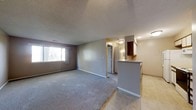 This single-floor space features an open-concept layout that seamlessly connects the living area and kitchen. The living room is finished with wall-to-wall carpeting in a neutral gray tone, complemented by light gray walls and white baseboards that create a clean, understated aesthetic. A window on the left wall allows natural light to filter into the space, offering a view of the surrounding greenery outside. The textured ceiling adds a classic touch throughout the unit.

Transitioning toward the right side of the space, the flooring shifts from carpet to tile, delineating the kitchen and dining area. A raised breakfast bar with a light-colored countertop extends from the kitchen, providing a casual dining or workspace option. Above the bar, dark-stained cabinetry is installed, offering ample overhead storage. The kitchen area is well-lit with built-in ceiling lighting.

A hallway visible toward the back of the space leads to what appears to be a bathroom, with a vanity and mirror visible through the doorway. The overall ambiance of the space is practical and functional, with neutral tones and straightforward finishes throughout.