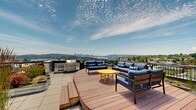 Located in Seattle's University District, this rooftop terrace at 4348 9th Ave NE offers an impressive outdoor amenity space with panoramic views of the Seattle skyline, Lake Union, and the greater Puget Sound region. The expansive rooftop is thoughtfully designed with multiple distinct zones to accommodate a variety of activities. Generous tiled patio areas are furnished with multiple high-top dining tables and bright yellow chairs, creating a vibrant and welcoming atmosphere for gatherings. A dedicated lounge zone features a warm wood deck fitted with chaise lounges, ideal for relaxing and taking in the views. The rooftop is also equipped with a stainless steel built-in grill and counter space, providing a fully functional outdoor kitchen for entertaining. A permanent ping-pong table adds a recreational element to the space. Throughout the terrace, galvanized metal planters filled with ornamental grasses and seasonal plantings provide greenery and visual interest. Elevator access serves the rooftop directly, ensuring convenient entry. The space offers unobstructed sightlines in multiple directions, encompassing downtown Seattle's high-rises, residential neighborhoods, and natural landscapes beyond.