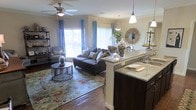 This single-floor residence offers an inviting open-concept design that seamlessly connects the living area and kitchen into one cohesive, functional space. The living room is anchored by rich hardwood flooring and a decorative area rug with a floral pattern, adding warmth and texture underfoot. Large windows dressed with light blue curtains flood the space with abundant natural light, creating a bright and airy atmosphere. A ceiling fan with an integrated light fixture adds both comfort and a casual charm to the room. The kitchen features dark cabinetry contrasted by granite countertops, a double sink, and a peninsula that opens directly to the living area — ideal for entertaining. A pendant light hangs above the peninsula, providing focused task lighting while contributing to the overall aesthetic. Decorative accents, including glass apothecary jars and a sunburst mirror, add character and a curated, welcoming feel throughout the space. A freestanding shelf unit along one wall offers display and storage opportunities. The overall ambiance is warm, comfortable, and thoughtfully styled, blending functional design with tasteful decor elements that make the space feel both livable and refined.