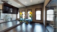 This fitness facility is thoughtfully divided into two distinct yet connected areas designed to support a wide range of exercise activities. The group exercise and yoga studio features rich, dark hardwood flooring that extends throughout, providing a warm yet polished foundation for movement-based workouts. Arched windows dressed with patterned roman shades allow natural light to filter in, brightening the warm taupe-toned walls. Full-length mirrors line one wall, amplifying the sense of space and allowing participants to monitor their form. A wall-mounted flat-screen television adds a multimedia element for guided sessions. A well-stocked equipment rack holds an assortment of stability balls, resistance bands, medicine balls, and rolled yoga mats, keeping essentials organized and accessible. Decorative yoga silhouette murals adorn the walls, reinforcing the studio's wellness-focused identity. Adjacent to this space, visible through a full glass partition, is a dedicated cardio and strength training area equipped with cycling bikes, treadmills, and weight machines. Recessed lighting throughout ensures consistent illumination across both spaces. Together, the two areas create a comprehensive, professional-grade fitness environment suited for individual training and group classes alike.