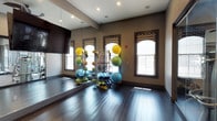 This fitness facility is thoughtfully divided into two distinct yet connected areas designed to support a wide range of exercise activities. The group exercise and yoga studio features rich, dark hardwood flooring that extends throughout, providing a warm yet polished foundation for movement-based workouts. Arched windows dressed with patterned roman shades allow natural light to filter in, brightening the warm taupe-toned walls. Full-length mirrors line one wall, amplifying the sense of space and allowing participants to monitor their form. A wall-mounted flat-screen television adds a multimedia element for guided sessions. A well-stocked equipment rack holds an assortment of stability balls, resistance bands, medicine balls, and rolled yoga mats, keeping essentials organized and accessible. Decorative yoga silhouette murals adorn the walls, reinforcing the studio's wellness-focused identity. Adjacent to this space, visible through a full glass partition, is a dedicated cardio and strength training area equipped with cycling bikes, treadmills, and weight machines. Recessed lighting throughout ensures consistent illumination across both spaces. Together, the two areas create a comprehensive, professional-grade fitness environment suited for individual training and group classes alike.