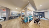 This thoughtfully designed single-floor amenity lounge offers a versatile and visually cohesive environment organized into several distinct functional zones. Light wood-look flooring runs throughout much of the space, complemented by decorative geometric tile insets that define specific areas and add visual interest underfoot.

A central lounge area anchors the space, featuring teal upholstered booth-style seating set against a slate-blue accent wall adorned with gold sculptural disc artwork. Pendant lighting and a built-in coffee station nearby enhance the social atmosphere. An adjacent bar-height counter provides additional casual seating beneath a recessed ceiling detail with warm wood paneling and a mounted television.

A dedicated game and recreation zone includes a full shuffleboard table along one wall, framed artwork, and direct access to an outdoor pool area through glass double doors. A separate media lounge features a large built-in entertainment wall with cabinetry, a mounted flat-screen TV, leather sofa, and additional seating, also opening to the outdoors.

A co-working or dining area accommodates group gatherings with long communal tables and pendant lighting, while wooden slatted dividers provide architectural definition between zones. Throughout, the design balances contemporary finishes with warm, inviting tones, creating an engaging and multifunctional communal environment.